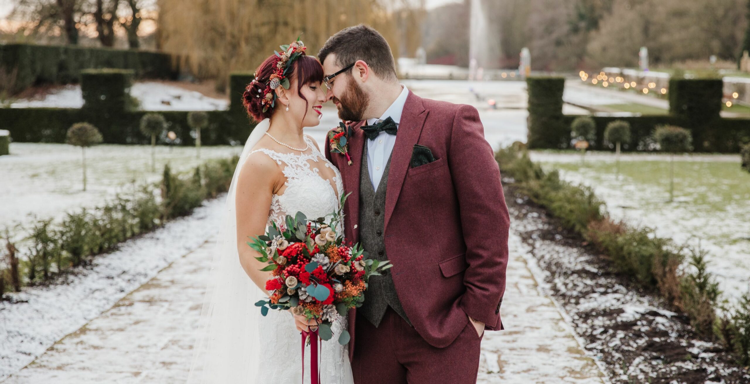Couple lovingly stands next to each other, bride holding bouquet and snow-covered Coombe Abbey estate provides perfect back drop, an affordable wedding venue